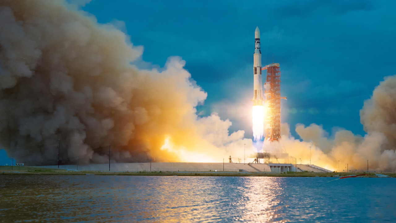 Rocket launching with bright flames against a blue sky background, large smoke clouds billowing near a calm water surface.