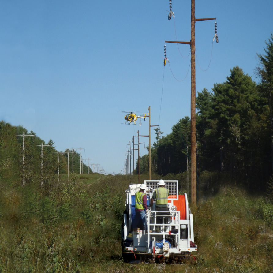 Utility workers in a vehicle inspect power lines as a helicopter hovers near them in a rural area with clear blue sky.