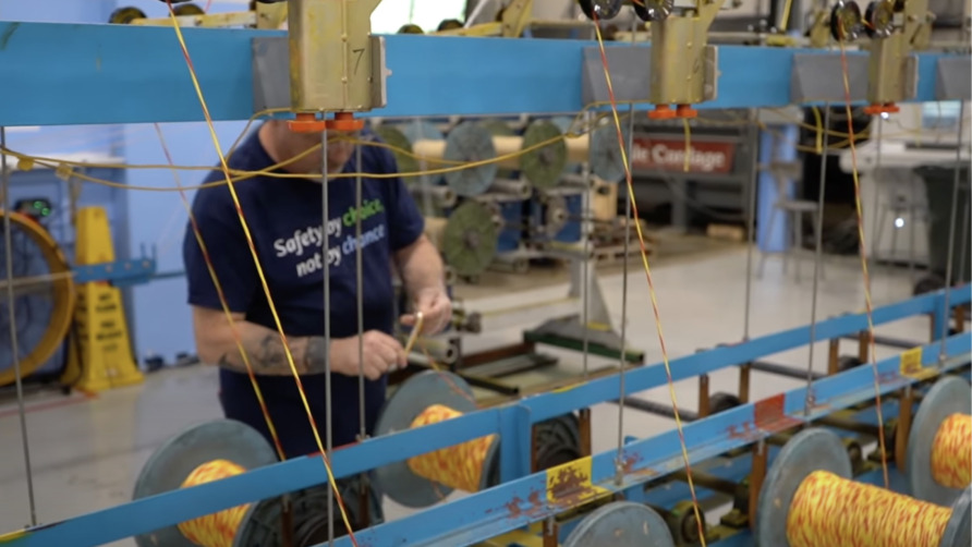 Worker operating machine in factory, spooling yellow and red thread onto large reels, with safety sign in background.