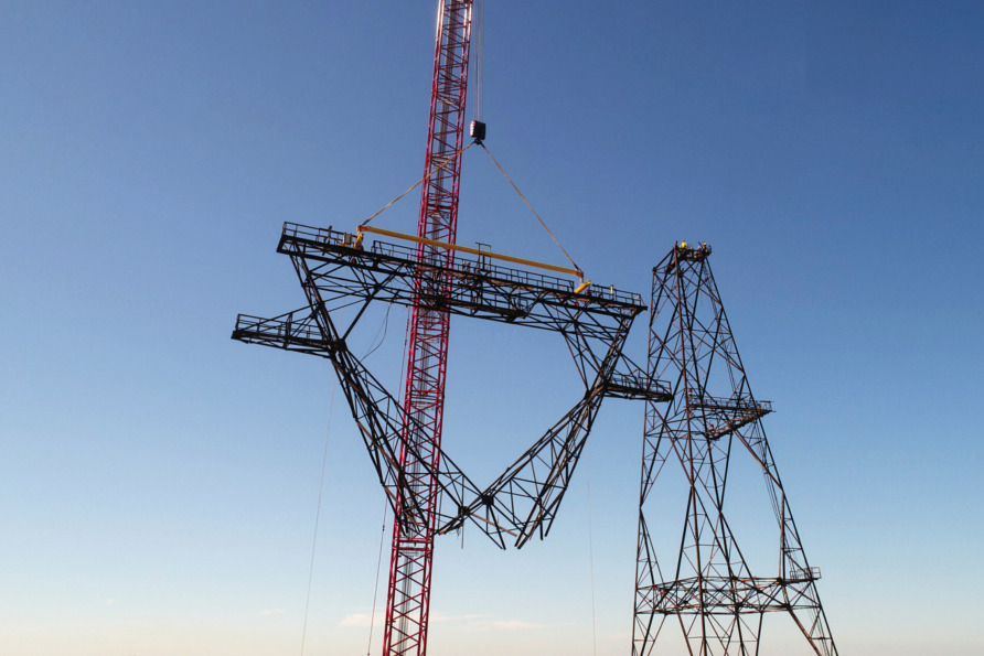 Crane lifting large steel pylon segment against clear blue sky during power line construction.