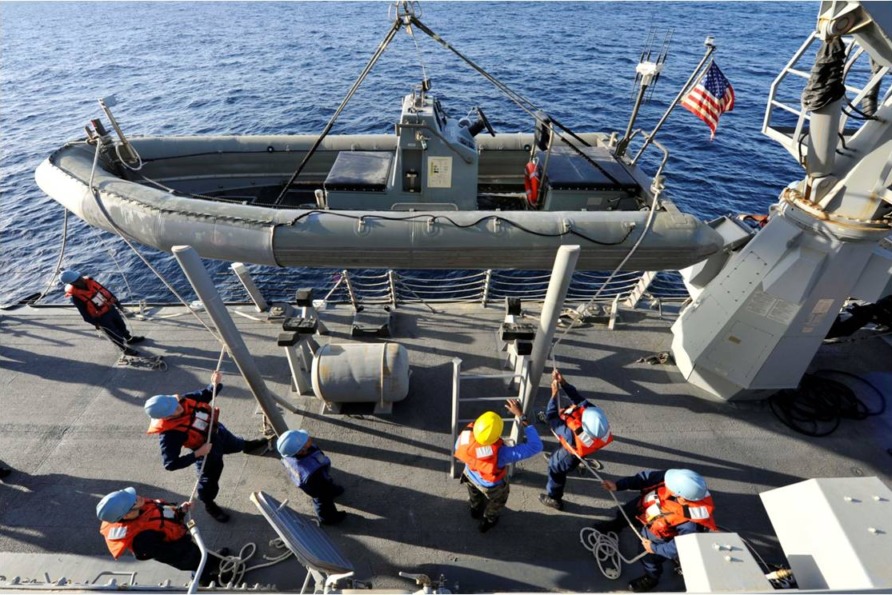 Crew members on a ship working together to hoist a lifeboat over the side, with an American flag visible in the background.