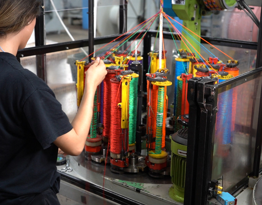 Person operating a colorful industrial braiding machine in a factory setting, focusing on spools of thread.