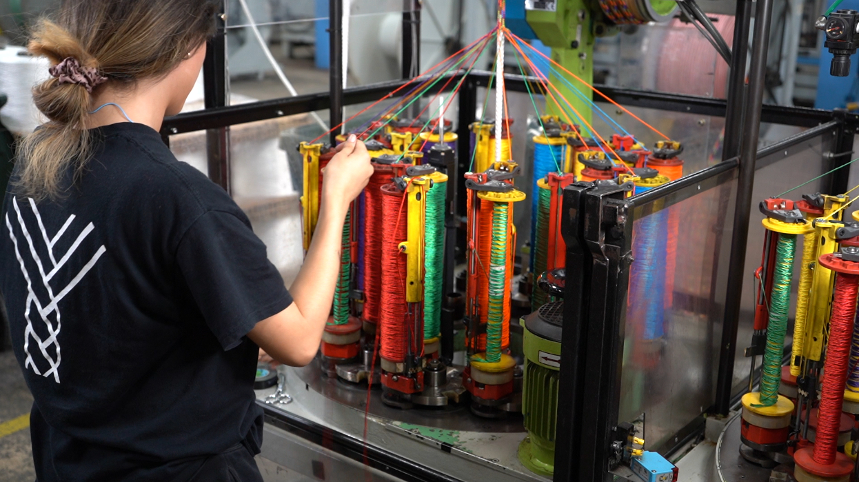 Person operating a colorful industrial braiding machine in a factory setting, focusing on spools of thread.