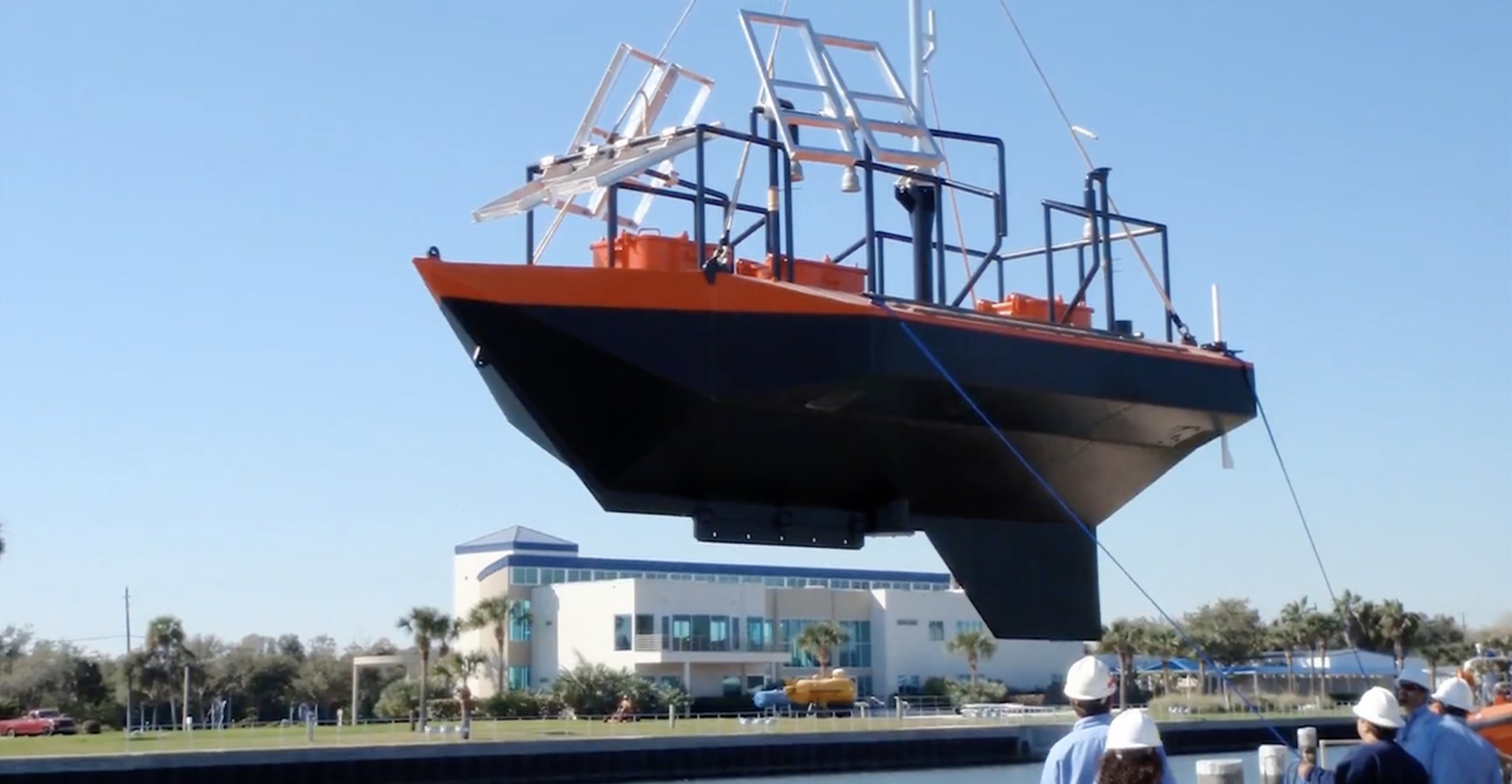 Boat suspended by crane over water, with people observing and modern building in the background.