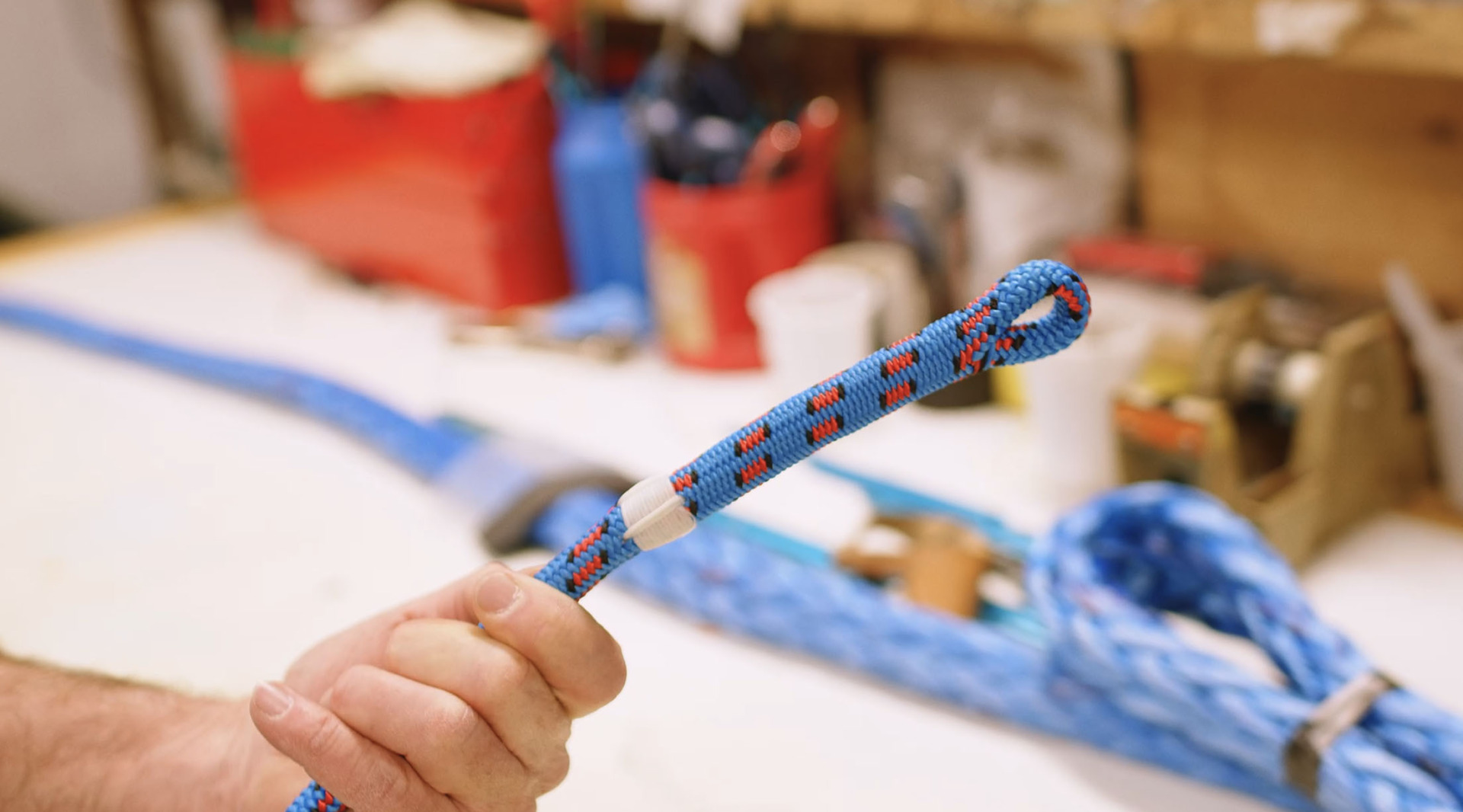 A hand holding a blue and red rope with a knot, workshop tools and materials blurred in the background.