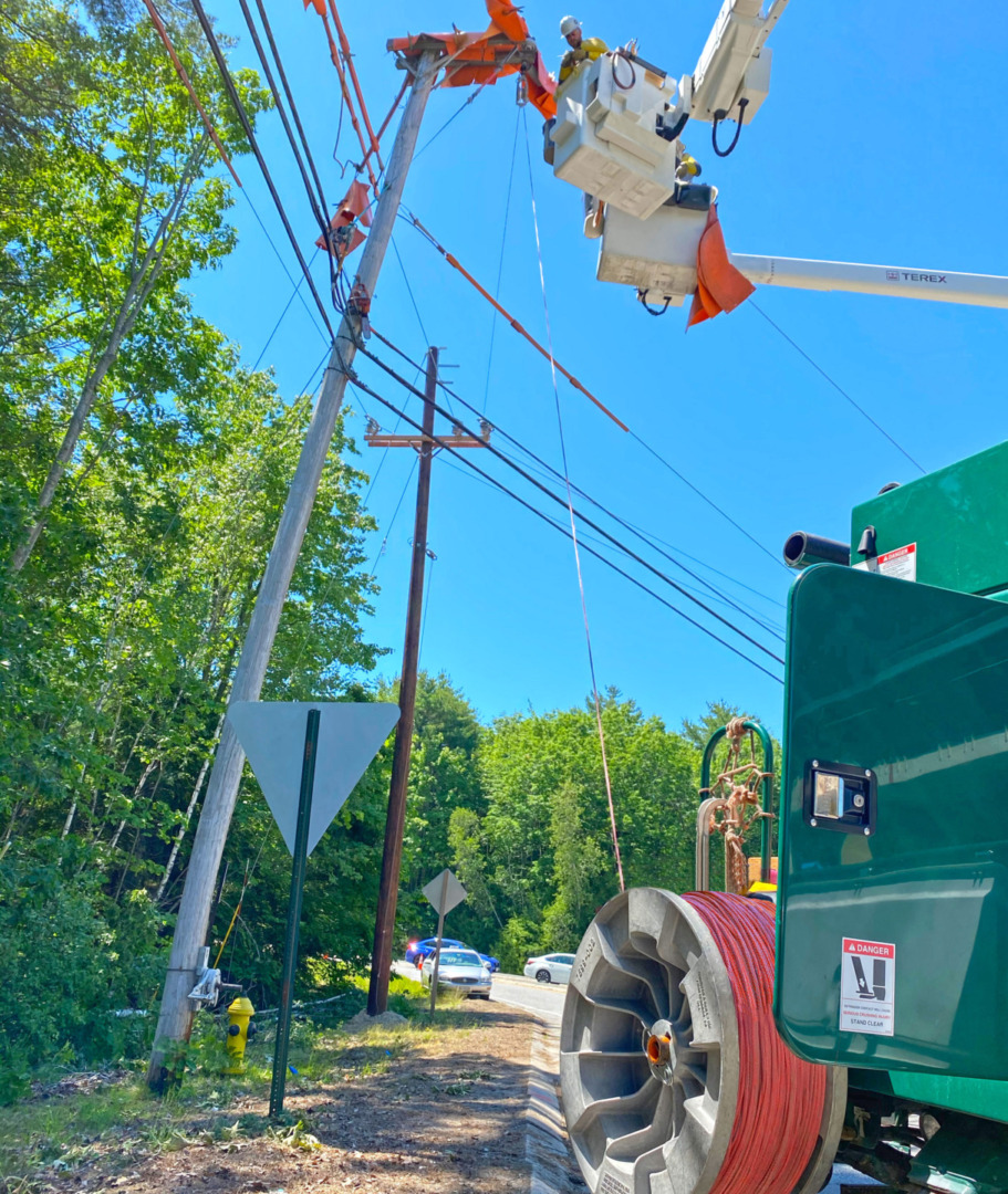 Utility workers in a bucket truck fixing power lines on a sunny day, surrounded by trees and blue sky.