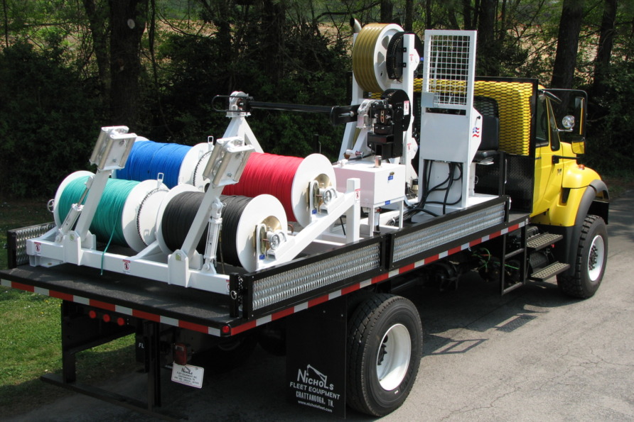 Truck with mounted cable reels in various colors parked on a road, surrounded by green trees.