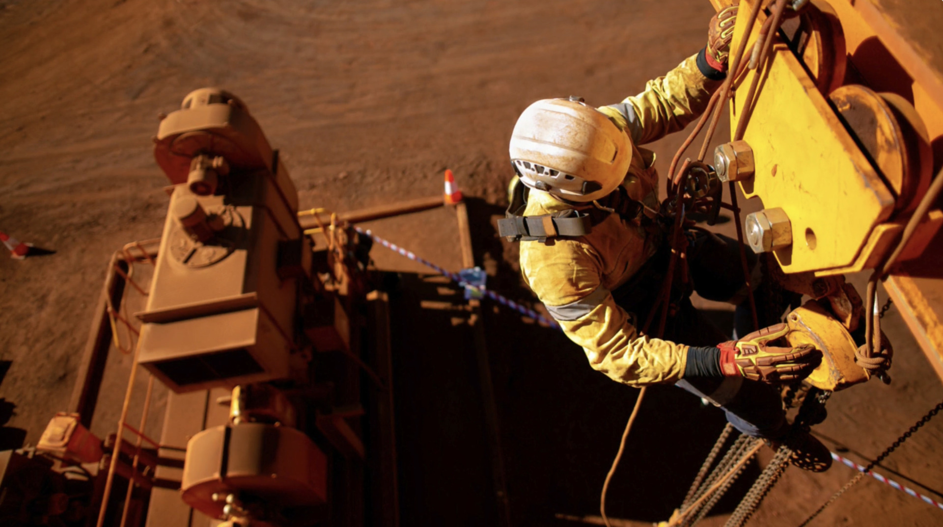 Worker in a safety harness climbs industrial equipment at a mining site, viewed from above with machinery in the background.