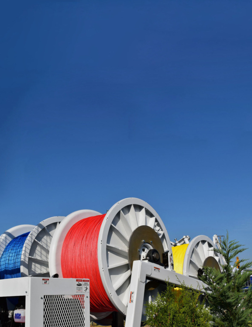Large industrial spools with red, blue, and yellow cables under a clear blue sky, surrounded by greenery.