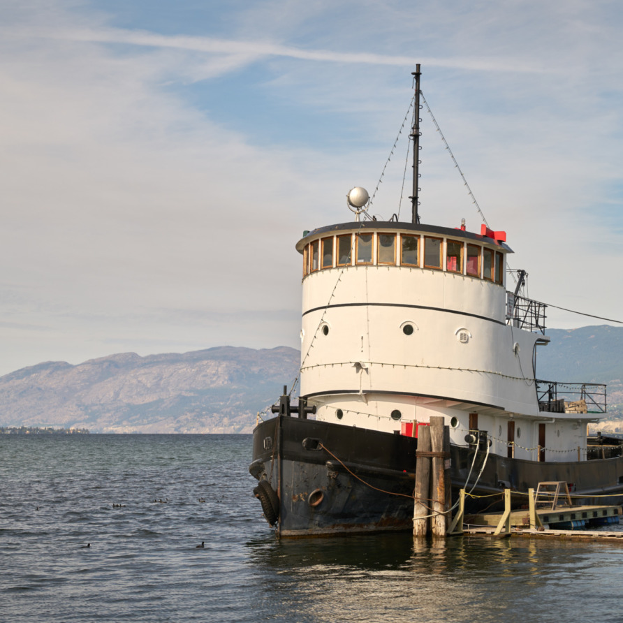 A vintage ferry docked on a calm lake with mountains in the background under a partly cloudy sky.