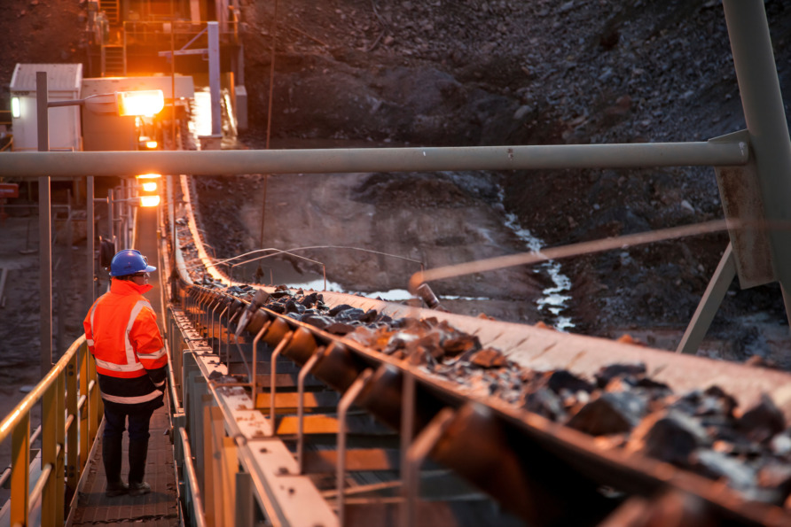 Worker in high-visibility gear observing a conveyor belt system at an industrial mining site during sunset.