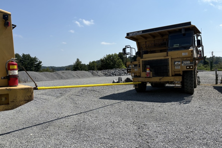 Large construction dump truck parked on gravel lot with caution tape, trees, and blue sky in the background.