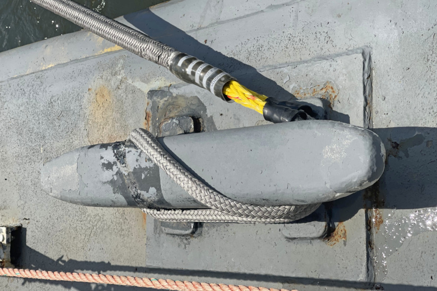 Rope tied around a gray bollard on a boat deck with a yellow and black cable nearby.