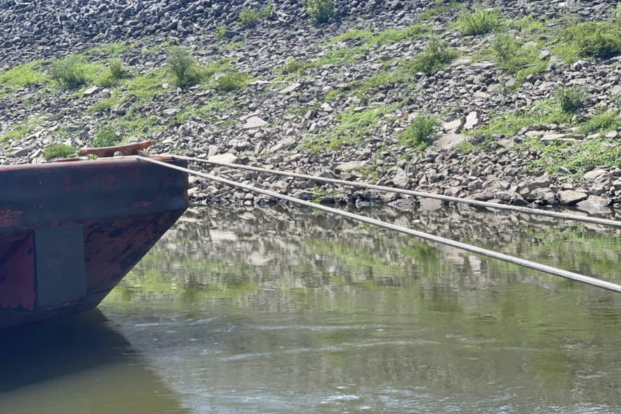 Rusty barge moored by ropes on calm waters near rocky shoreline with green vegetation.