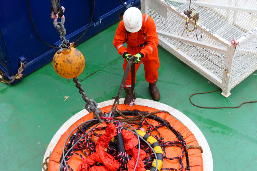 Worker in orange safety gear securing cargo with straps on a ship deck, using safety hoist equipment.