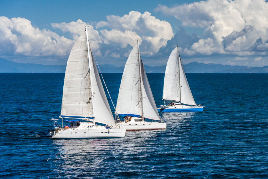 Three sailboats with white sails gliding on blue ocean water under a partly cloudy sky