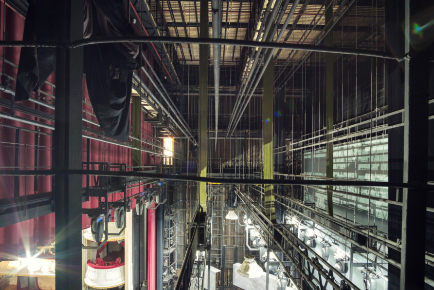 View of a theater stage from backstage, showing rigging, curtains, and stage lights in a detailed architectural setting.