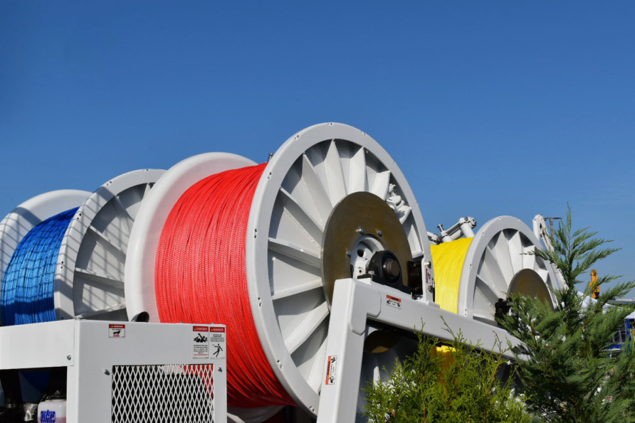 Large spools of colorful cables in blue, red, yellow, and white under a clear blue sky, surrounded by greenery.