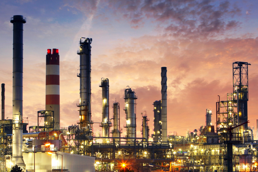 Industrial refinery with smoke stacks against a sunset sky, featuring a large red and white striped chimney and various structures.