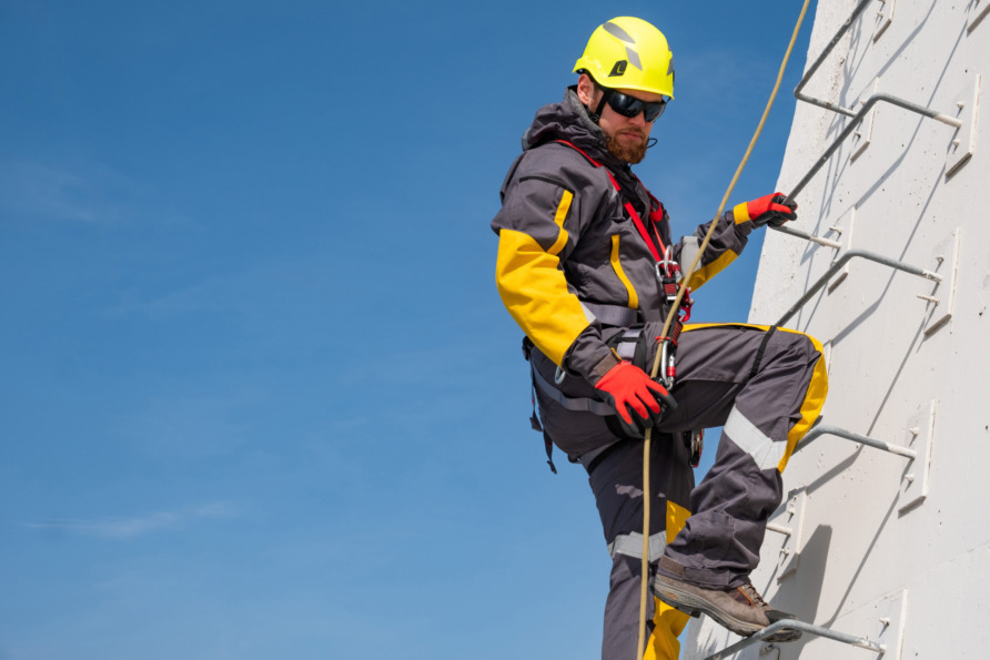 Worker in safety gear climbing a ladder on a wind turbine against a clear blue sky.