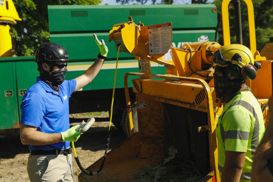 Workers operating a wood chipper, wearing protective gear, with a green truck in the background.