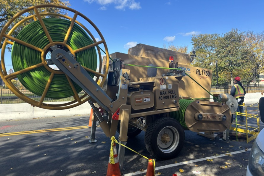Large machine with green coiled cable on a street, surrounded by cones and safety barriers.