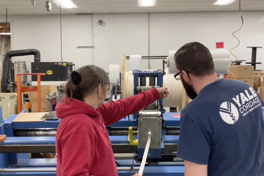 Two people examining a machine in a factory setting, one pointing towards a component, surrounded by equipment.