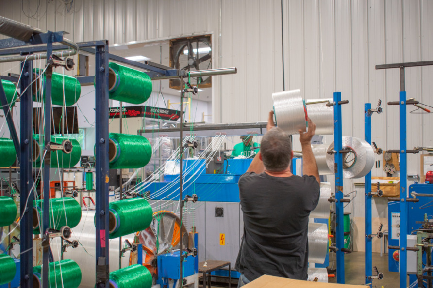 Worker in a factory handling spools of thread, surrounded by machinery and green thread rolls on racks.