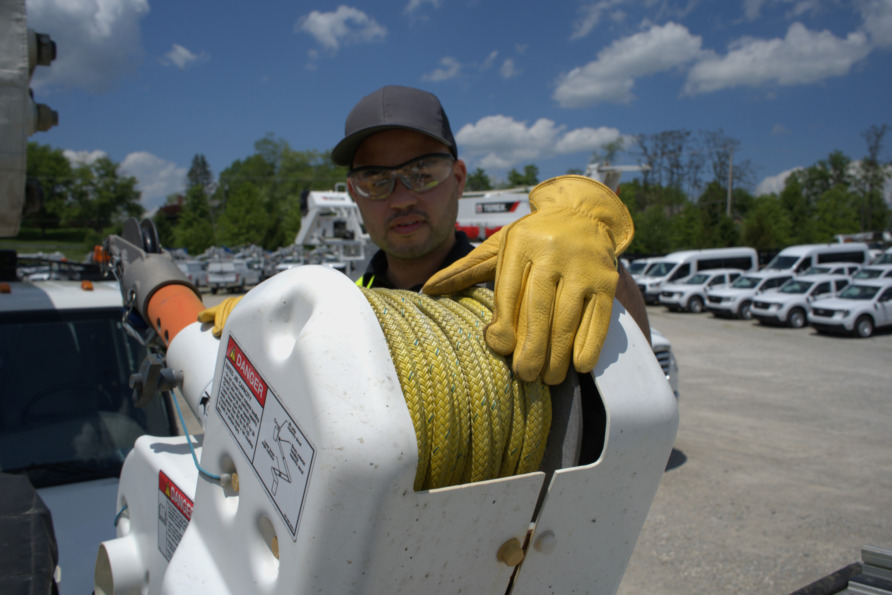 Worker in protective gear handling a yellow rope on a utility truck, parked with other service vehicles under a clear sky.