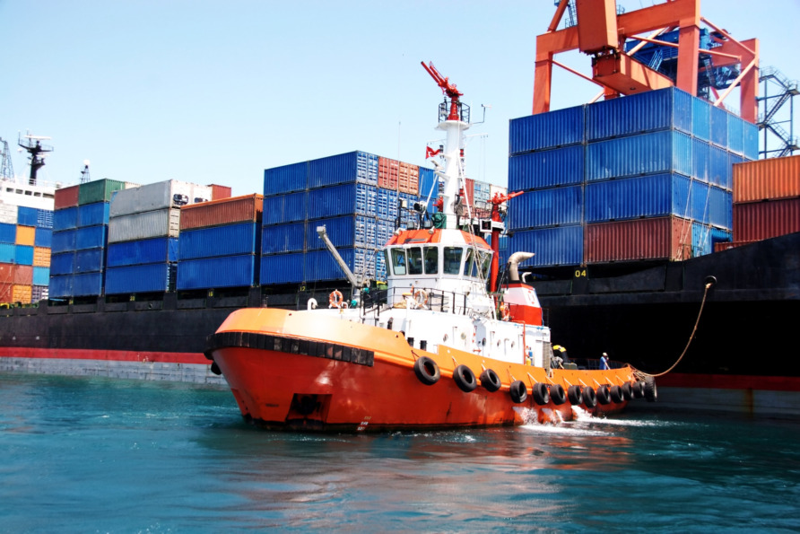 Tugboat guiding cargo ship loaded with colorful shipping containers in port under clear blue sky.