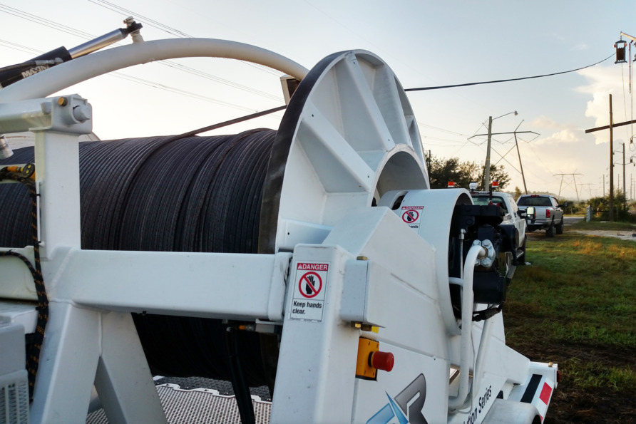 Large cable spool mounted on a truck, positioned on a roadside with power lines and vehicles visible in the background.