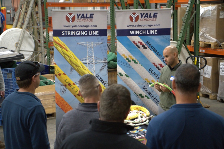Man presenting rope products to a group in a warehouse setting with "Yale Cordage" banners displayed behind him.