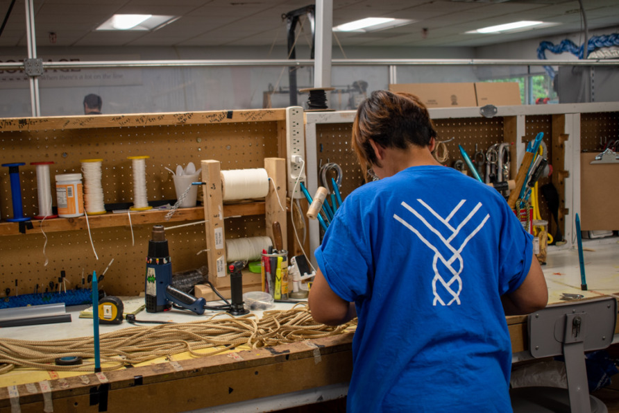 Person working at a workstation with tools, threads, and ropes in a workshop, wearing a blue shirt with a white design.