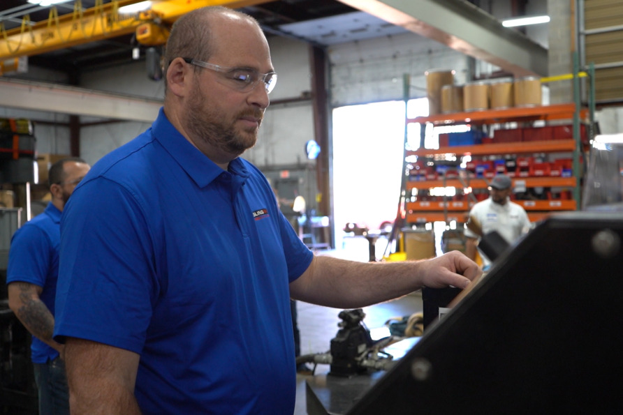 Man wearing safety glasses operates machinery in an industrial setting with shelves and equipment visible in the background.
