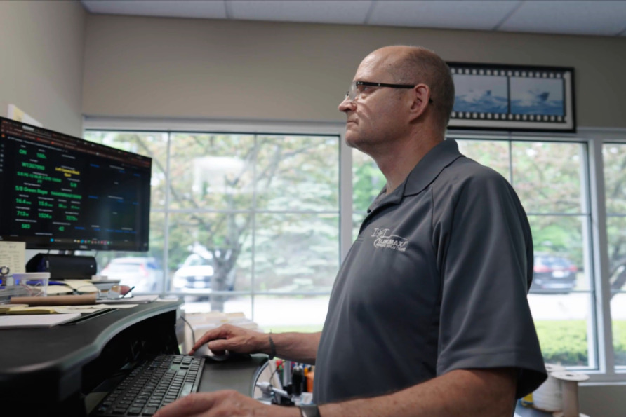 Man working at a computer desk in an office with large windows and a view of trees outside.
