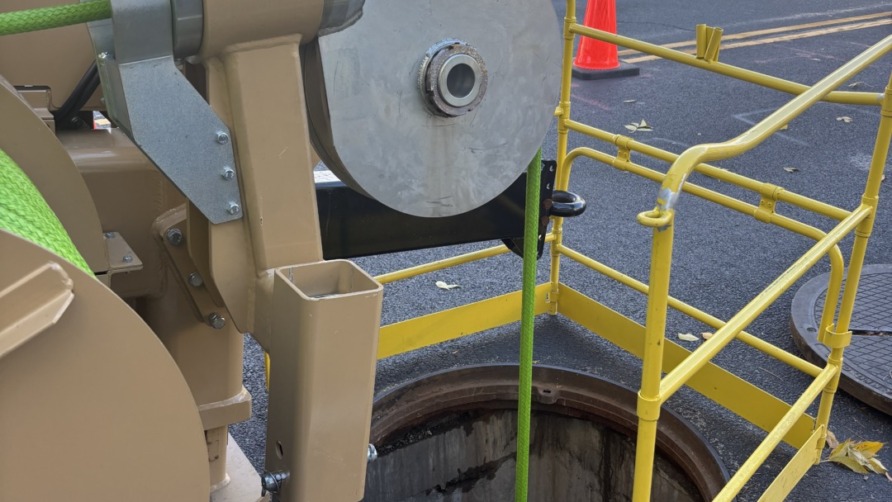 Winch system with metal cable and yellow guard rail at an open manhole on a street with autumn trees and fallen leaves.