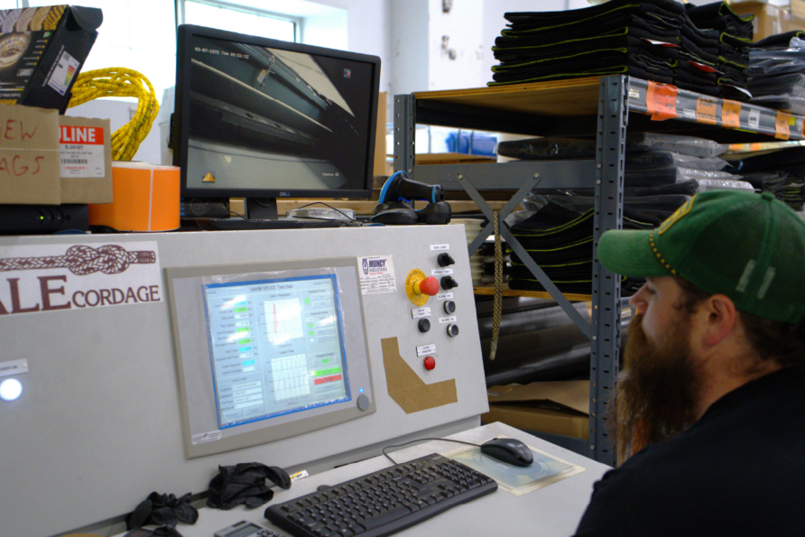 Man operating machinery in a manufacturing facility with computer screens displaying data and shelves in the background.