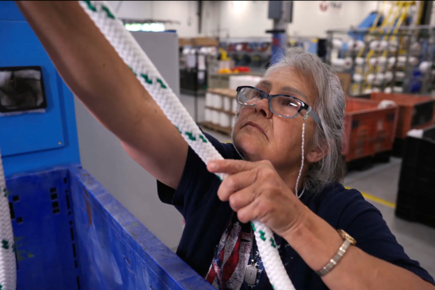 Woman with gray hair and glasses inspecting a braided rope in a factory setting.