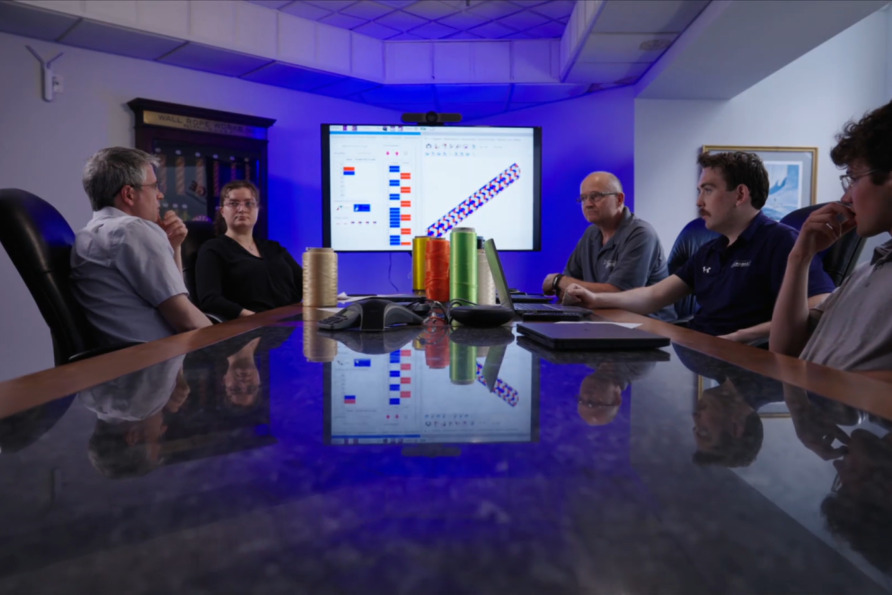 Team discussing data charts on screen in conference room with reflection on table, spools of thread on the side.