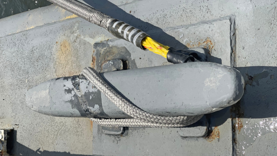 Rope tightly wrapped around a weathered metal bollard on a pier, securing a boat with a visible water surface beside.
